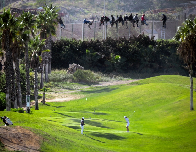 A golfer hits a tee shot as African migrants sit atop a border fence during an attempt to cross into Spanish territories between Morocco and Spain's north African enclave of Melilla